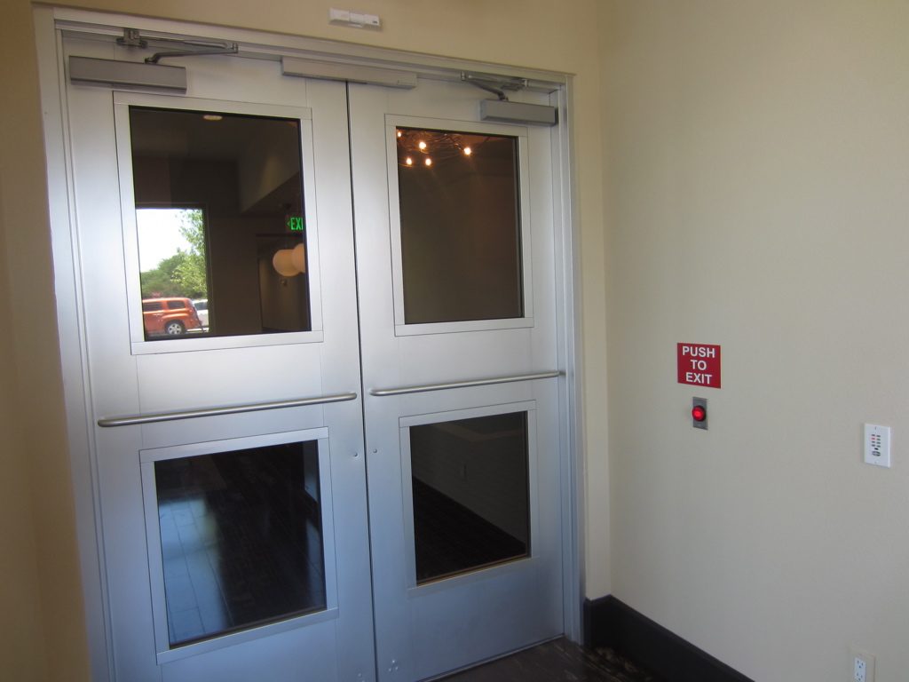 Pair of silver double doors with door closers and push bars, with a red PUSH TO EXIT sign and button on the wall beside them at a Dallas apartment building