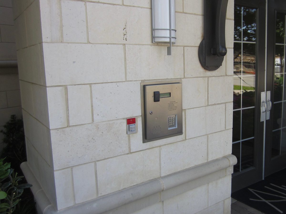 Stainless steel telephone entry panel with card reader, keypad and directory display set into a stone wall beside glass double doors at a Dallas apartment building