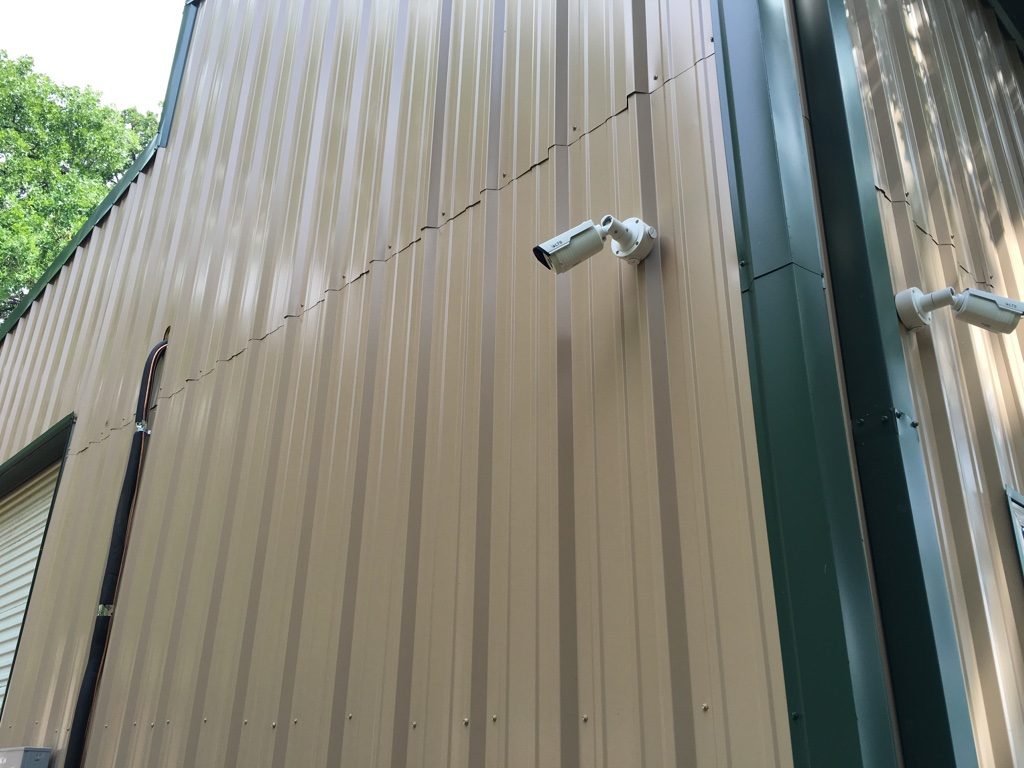 Two white bullet security cameras mounted on the tan metal siding of a barndominium with green trim