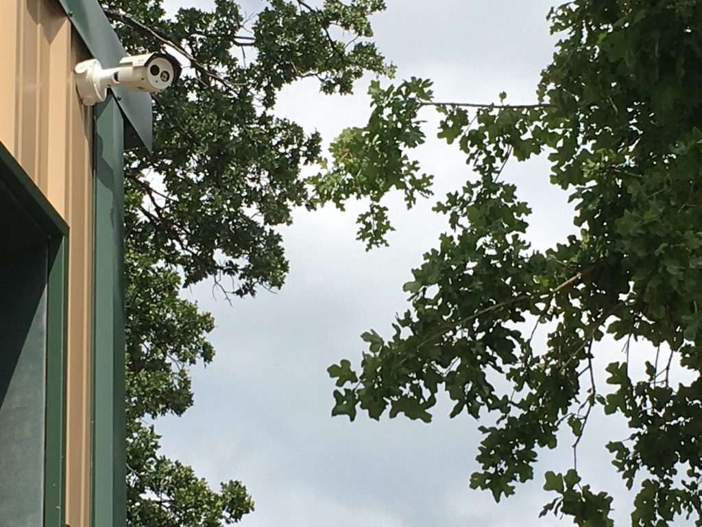 White bullet security camera mounted under the roofline of a barndominium with oak trees and sky behind it