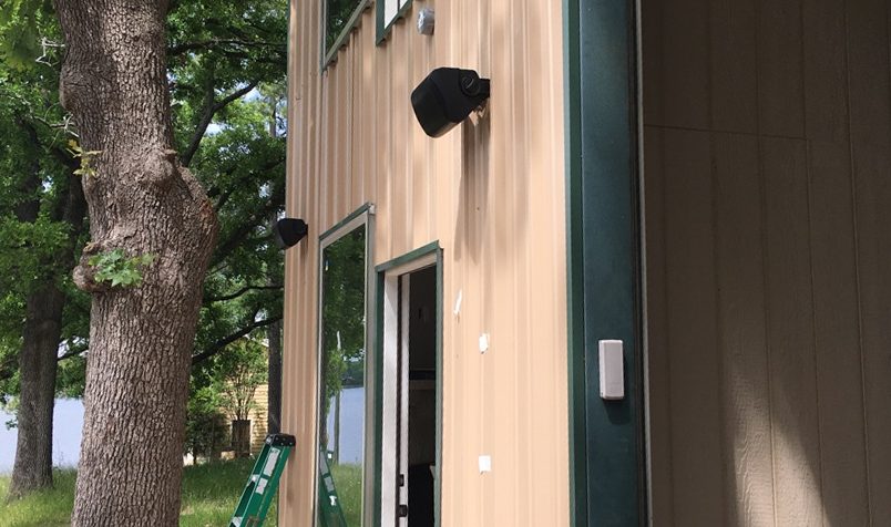 Black outdoor speakers mounted on the tan metal siding of a barndominium beside a door, with oak trees and a lake behind