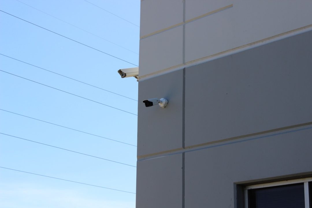 Two white bullet security cameras mounted on the corner of a large warehouse wall against a clear sky