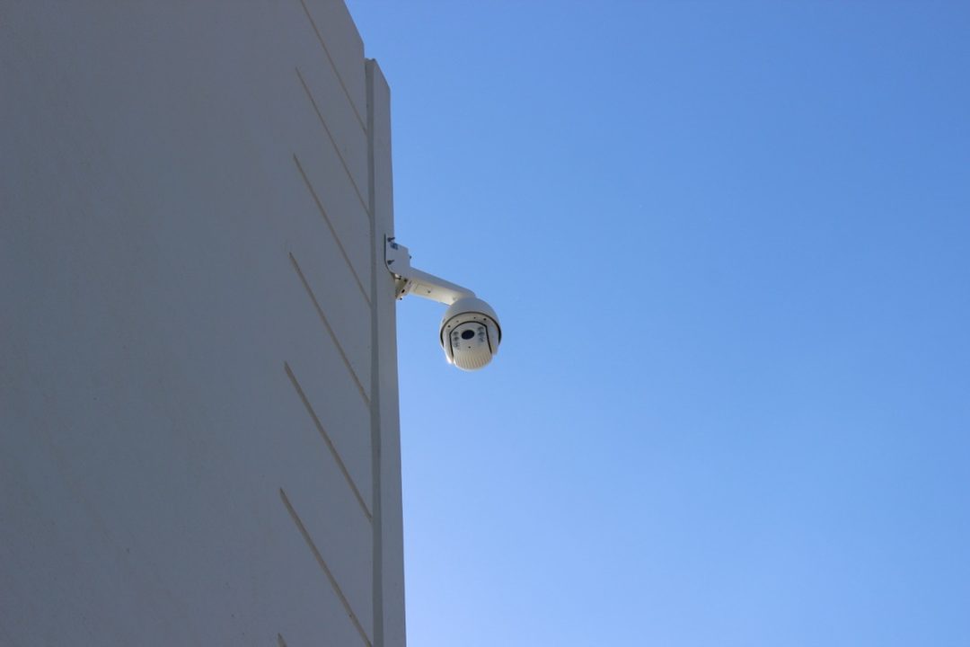 White dome security camera on a wall-mount arm extending from a warehouse wall against a blue sky