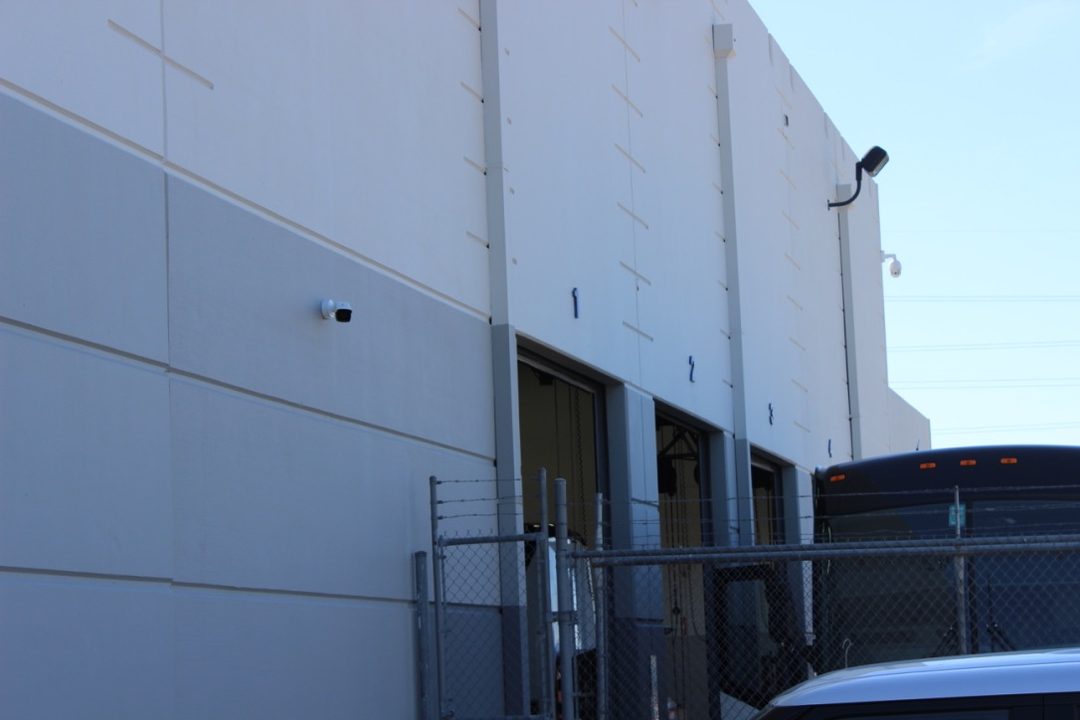 Warehouse wall with numbered loading dock doors, with a security camera on the wall and another camera and light near the roofline